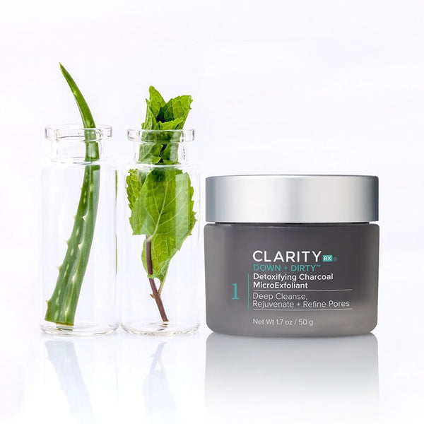 Jar of Down + Dirty™ Detoxifying Charcoal MicroExfoliant | ClarityRx next to glass containers holding aloe vera and green leaves, representing natural skincare ingredients.