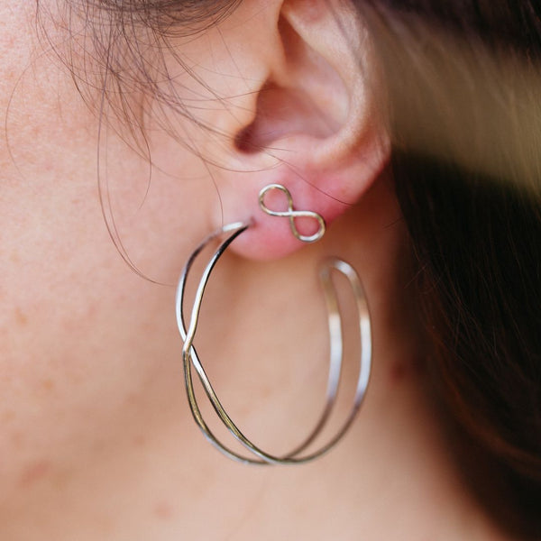 Close-up of a person wearing Infinity Hoops | Purpose Jewelry silver earrings featuring a small infinity symbol stud and a larger open hoop design, showcasing elegant minimalism.