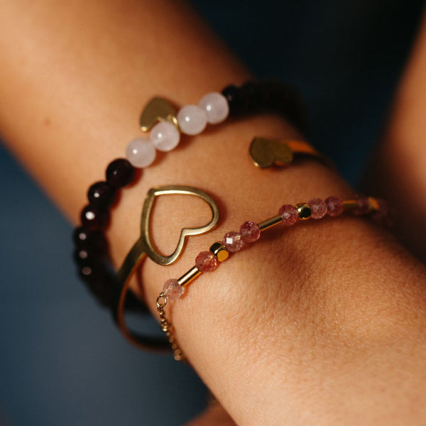 Close-up of a wrist adorned with multiple delicate bracelets featuring heart-shaped gold accents and gemstones, showcasing the Shine Bracelet | Purpose Jewelry.