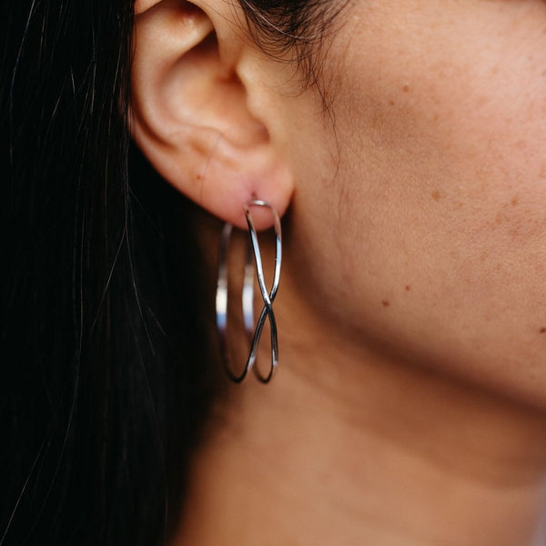 Close-up of a person wearing silver twisted infinity hoop earrings featuring a modern design, showcasing Infinity Hoops | Purpose Jewelry.