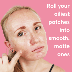 Close-up of a woman with oily skin pointing to her cheek against a pink background with text promoting the Slick Oil Absorbing Volcanic Roller [Removes Oil & Shine] | Dreambox Beauty for smoothing and mattifying oily patches.