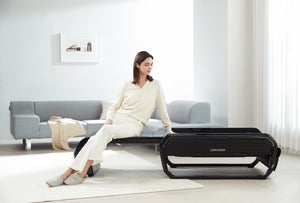 Woman in white loungewear sitting on the CERAGEM V6 Therapeutic Thermal Home Massager | Ceragem in a bright, minimalist living room setting.