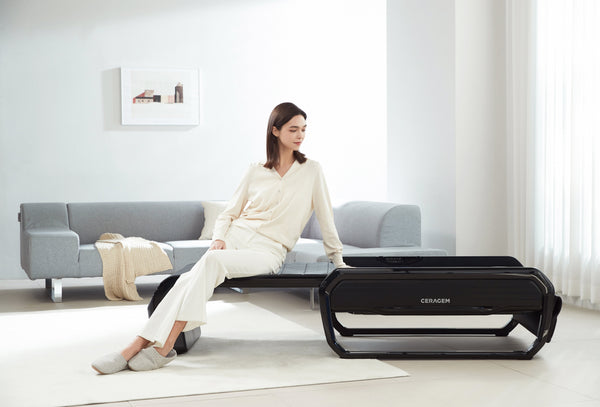 Woman in white loungewear sitting on the CERAGEM V6 Therapeutic Thermal Home Massager | Ceragem in a bright, minimalist living room setting.