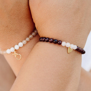 Close-up of two wrists wearing Gemstone Heart Bracelet | Purpose Jewelry featuring alternating dark and light gemstone beads with small gold heart and XO charms on a natural skin tone background.
