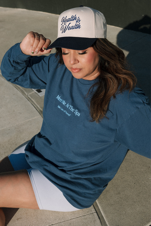 Woman wearing a navy blue Meet Me At The Spa - Take Care Of Yourself Long Sleeve T-Shirt | Lucky Owl sitting outdoors, paired with a white and navy cap that reads Healthy is Wealth.