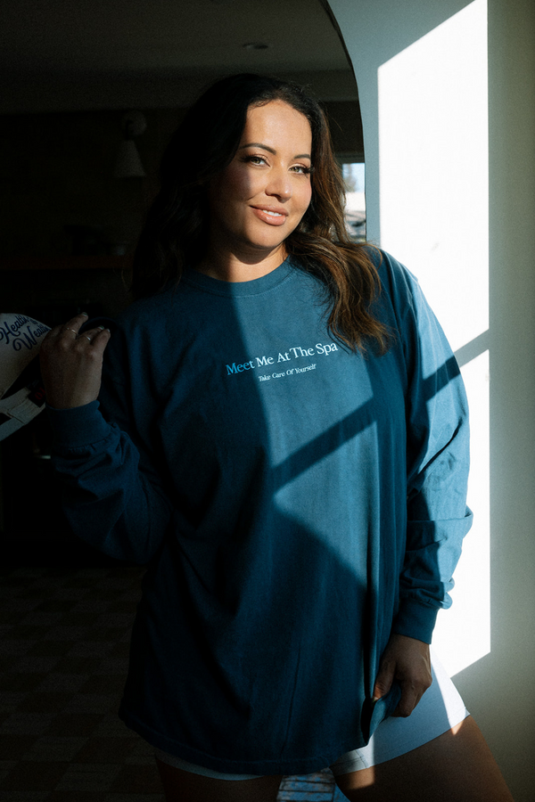 Woman wearing a blue "Meet Me At The Spa - Take Care Of Yourself Long Sleeve T-Shirt | Lucky Owl" standing in soft natural light near a window, promoting wellness and self-care.