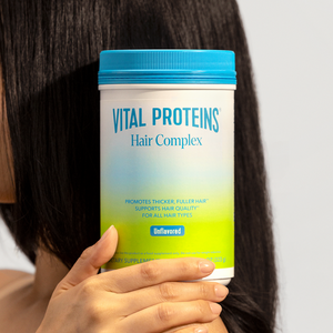 Person holding a container of Hair Complex - Unflavored | Vital Proteins supplement with long, healthy dark hair visible in the background promoting hair wellness and quality.
