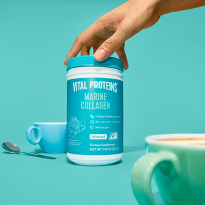 Person's hand holding a container of Marine Collagen - Unflavored | Vital Proteins next to a blue cup and spoon on a teal surface, highlighting wellness supplements for skin and joint support.