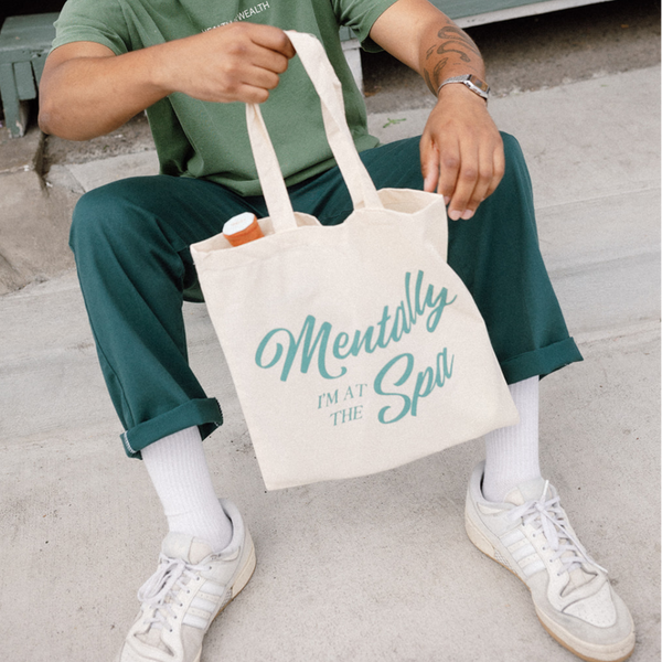 Person sitting outdoors holding a cotton canvas tote bag with the text "Mentally At The Spa Tote | Lucky Owl" containing wellness products, promoting relaxation and self-care.