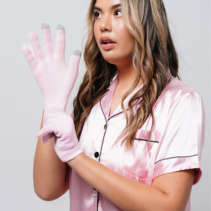 A woman wearing light pink Hydrating Infused Moisturizing Gloves | Dreambox Beauty over her hands, dressed in a matching pink satin pajama top, showcasing skincare and body care wellness.