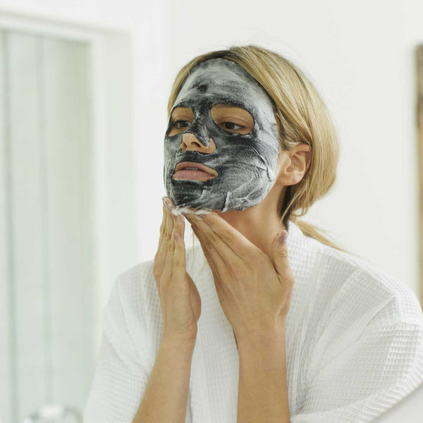 Woman wearing a black skincare sheet mask applying the 7-Minute Makeover Mask | Skin Authority in a bright bathroom, promoting skincare and wellness.