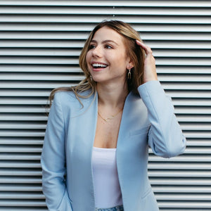 Woman wearing a light blue blazer and white top smiling while showing gold Infinity Hoops | Purpose Jewelry earrings against a modern striped background