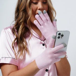Woman wearing pink Hydrating Infused Moisturizing Gloves | Dreambox Beauty with gray fingertips using a smartphone, demonstrating skincare and touchscreen compatibility.