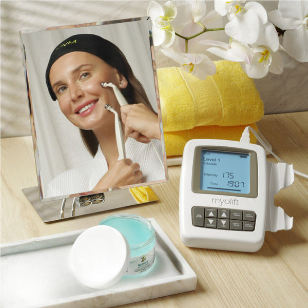 Woman using a facial wellness device in front of a mirror with a jar of ReStore Conductive Gel with Bio-Active Complex | 7e Wellness and yellow towels in the background