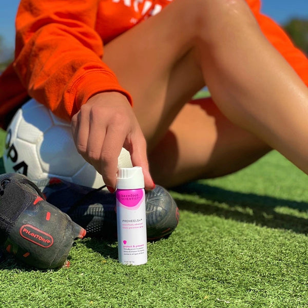 Person sitting on grass field near soccer cleats holding a bottle of Preheels+ Blister Prevention Spray | Barefoot Scientist with a soccer ball nearby, promoting skincare and body care for athletes.