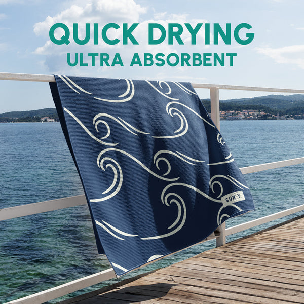 Waffle Microfiber Beach Towel - Curl | SUN'Y hanging on a wooden railing by the sea, featuring a navy blue color with white wave patterns, highlighting its quick drying and ultra absorbent qualities.