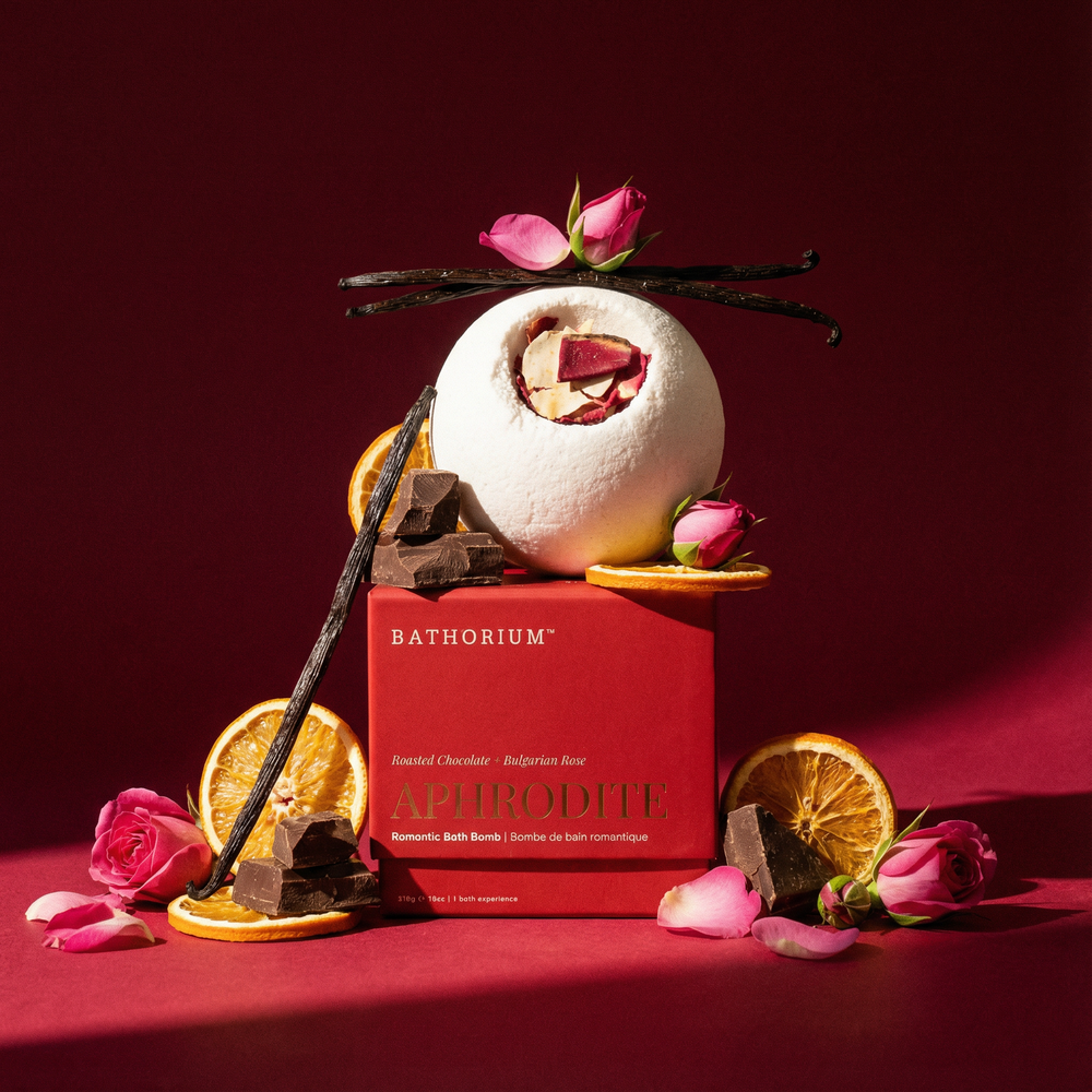 A white round bath bomb with dried rose petals on top displayed next to its red box packaging labeled Aphrodite Bath Bomb | Bathorium, featuring roasted chocolate and Bulgarian rose scents.