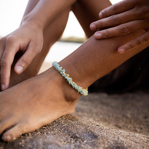 Close-up of a person's ankle wearing a delicate blue and gold beaded Fringe Bracelet | Catherine Weitzman Jewelry resting on sandy ground outdoors.
