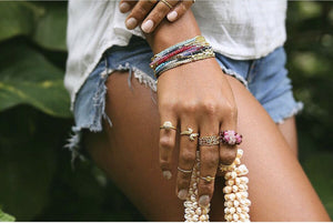 Close-up of a hand wearing multiple colorful bracelets and rings, including the Gemstone Fade Bracelet | Catherine Weitzman Jewelry, holding a strand of white shells against a casual outfit background.