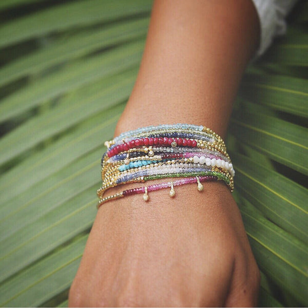 Close-up of a wrist wearing the Gemstone Fade Bracelet | Catherine Weitzman Jewelry, featuring multiple delicate strands with colorful gemstones and gold beads against a green leaf background.