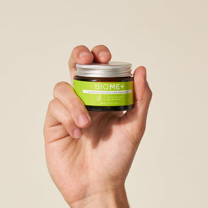 Hand holding a small jar of BIOME+ smoothing cloud crème | IMAGE Skincare with a green label against a neutral background, highlighting skincare and wellness.