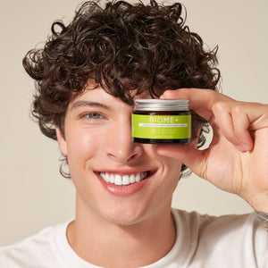 Young person with curly hair smiles holding a jar of BIOME+ smoothing cloud crème | IMAGE Skincare close to their eye, promoting skincare and wellness products.