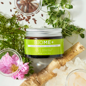 Jar of BIOME+ smoothing cloud crème | IMAGE Skincare on a white surface surrounded by natural elements including green leaves, a pink flower, glass bowls, and a piece of root, promoting skincare and wellness.