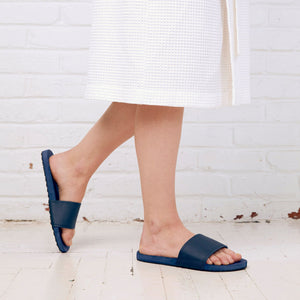 Person wearing navy Spa/Shower Slide | Feelgoodz slippers with a textured sole, standing on a white wooden floor against a white brick wall.