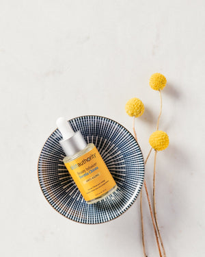 Beauty Infusion Swertia Chirata for Anti-Aging | Skin Authority serum bottle with dropper in a decorative blue and white bowl beside three yellow billy ball flowers on a light marble surface.