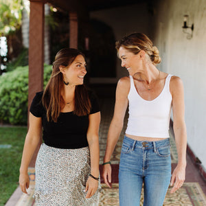 Two women smiling and walking outdoors together, one wearing a black top and floral skirt with the Dusk Stretch Bracelet | Purpose Jewelry on her wrist, enjoying a casual moment of connection.