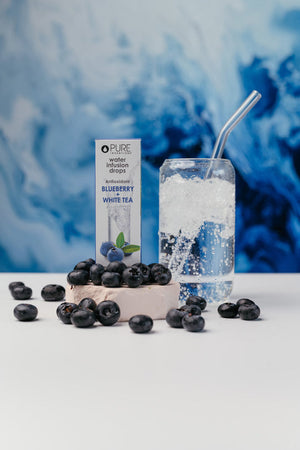Super Fruit Extracts Water Infusion Drops - Blueberry + White Tea | Pure Inventions displayed next to fresh blueberries and a glass of sparkling water with a metal straw on a white surface with a blue abstract background.