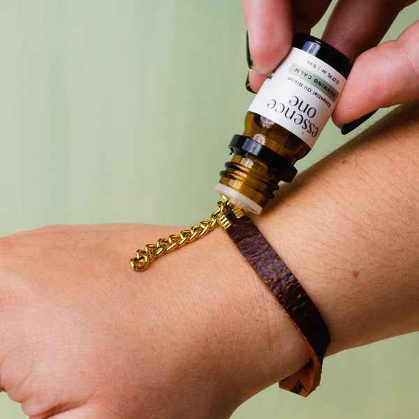 Hand applying Essential Oil Blends | Essence One onto a brown leather bracelet with a gold chain, highlighting a wellness and aromatherapy moment.