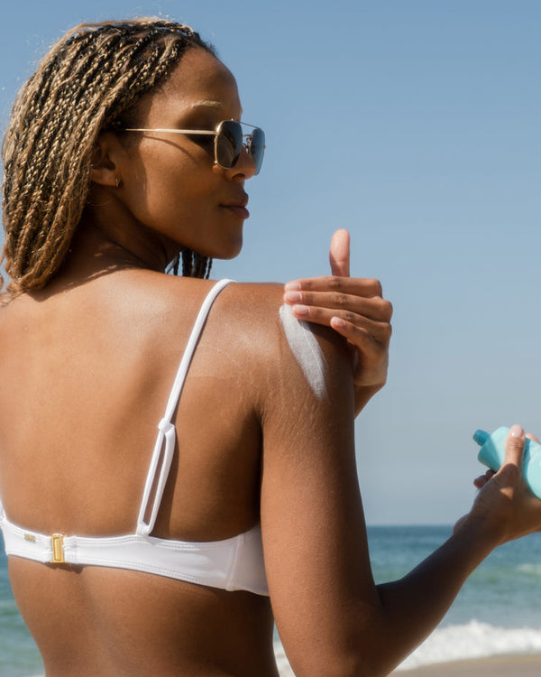 Woman wearing sunglasses and a white bikini top applies Peach Blossom Classic Body Sunscreen Lotion SPF 70 | COOLA to her shoulder at the beach with the ocean in the background.