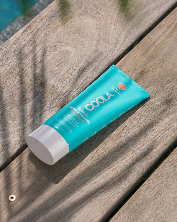 Peach Blossom Classic Body Sunscreen Lotion SPF 70 | COOLA in a turquoise tube resting on a wooden surface with soft shadows, highlighting skincare and sun protection.