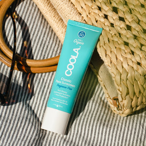 Classic Face Organic Sunscreen Lotion SPF 50 - Fragrance Free | COOLA tube placed on striped fabric next to woven straw bag and round wooden sunglasses, showcasing skincare essentials for sun protection.