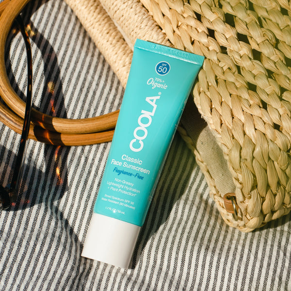 Classic Face Organic Sunscreen Lotion SPF 50 - Fragrance Free | COOLA tube placed on striped fabric next to woven straw bag and round wooden sunglasses, showcasing skincare essentials for sun protection.