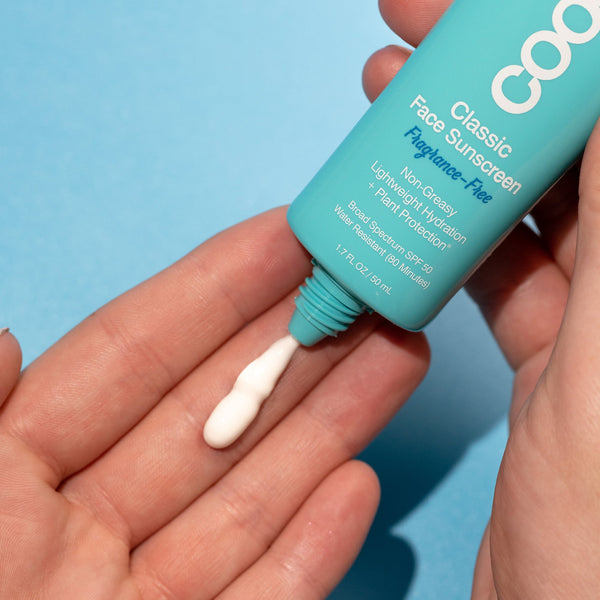 A person dispensing creamy white sunscreen lotion onto their fingertips from a turquoise tube of Classic Face Organic Sunscreen Lotion SPF 50 - Fragrance Free | COOLA against a light blue background.