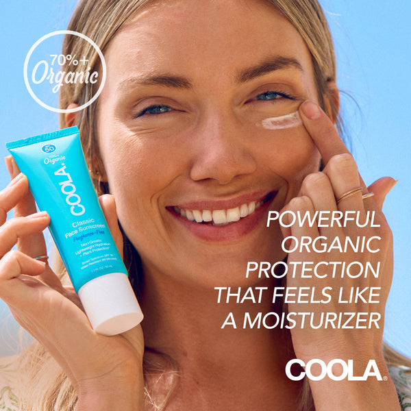 Smiling woman applying Classic Face Organic Sunscreen Lotion SPF 50 - Fragrance Free | COOLA to her face, highlighting skincare and organic protection with a bright blue sky background.