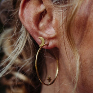Close-up of an ear wearing Galaxy Studs | Purpose Jewelry, featuring a gold hoop earring with a crescent moon at the top and a star charm hanging inside the hoop.