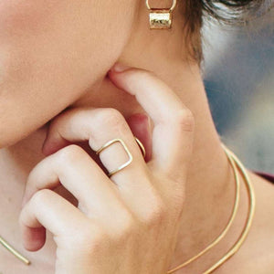 A close-up of a hand wearing a delicate, modern gold geometric ring, styled with a minimalist gold necklace and hoop earring, showcasing the Chloe Ring | Purpose Jewelry.