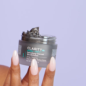 Hand holding an open jar of Down + Dirty™ Detoxifying Charcoal MicroExfoliant | ClarityRx showing the dark textured exfoliant inside against a light purple background