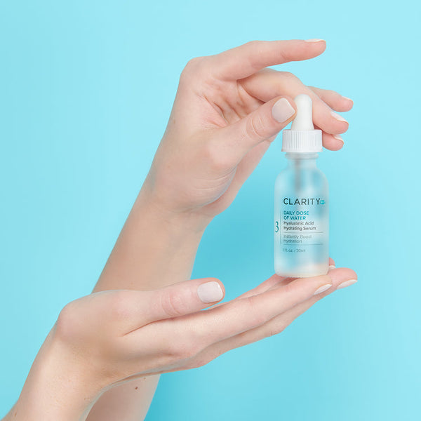 A pair of hands holding a frosted glass dropper bottle of Daily Dose Of Water™ Hyaluronic Acid Hydrating Serum | ClarityRx against a light blue background, showcasing skincare product and hydration benefits.