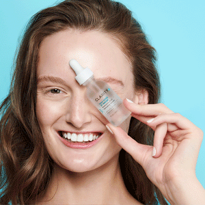 Smiling woman with clear skin holding a small bottle of Daily Dose Of Water™ Hyaluronic Acid Hydrating Serum | ClarityRx against a light blue background, showcasing skincare hydration.