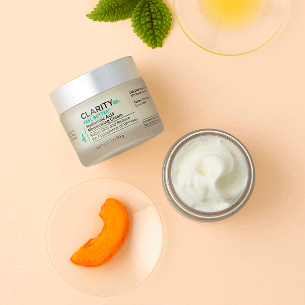 Open jar and closed container of Feel Better™ Hyaluronic Acid Moisturizing Cream | ClarityRx shown with fresh leaf, slice of orange fruit, and bowl of oil on beige background, highlighting skincare and hydration.