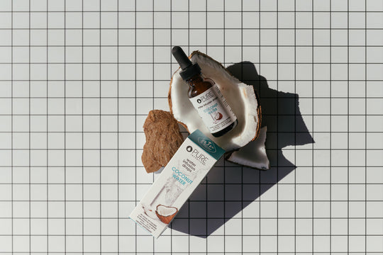 Coconut Water Infusion Drops | Pure Inventions bottle with dropper placed inside a halved coconut shell next to the product box on a grid-patterned surface, promoting wellness supplements.