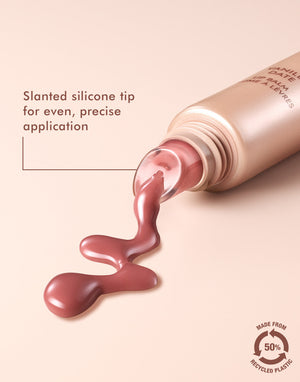 Close-up of Argan Butter Lip Balm | Moroccanoil with pinkish balm flowing from a tube featuring a slanted silicone tip for precise application, against a soft beige background.