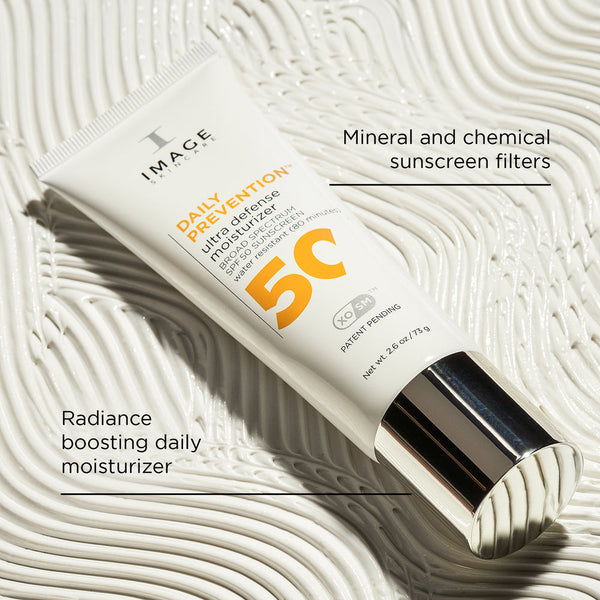 Tube of DAILY PREVENTION Ultra Defense Moisturizer SPF 50 | IMAGE Skincare with a silver cap placed on textured white background highlighting its radiance boosting and mineral and chemical sunscreen features.