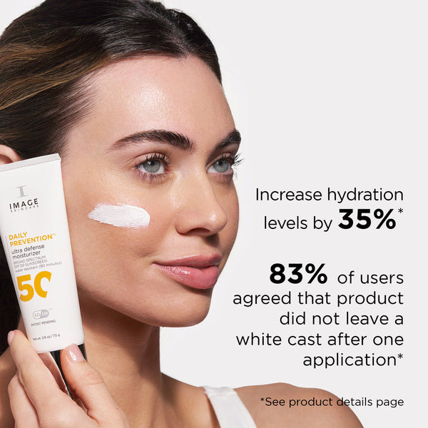 Woman holding DAILY PREVENTION Ultra Defense Moisturizer SPF 50 | IMAGE Skincare with a white cream swatch on her cheek, highlighting hydration and no white cast claims.