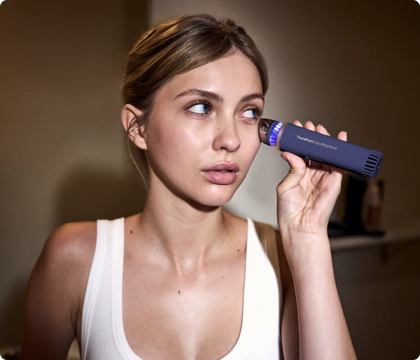 A woman using the TheraFace Depuffing Wand | Therabody with an indigo body on her under-eye area as part of a skincare routine for reducing puffiness and promoting wellness.
