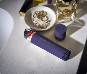TheraFace Depuffing Wand | Therabody in indigo color placed on a countertop near jewelry and a perfume bottle, representing a skincare wellness device.